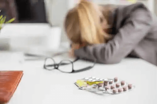 En kvinna vilar huvudet på sitt skrivbord, med olika mediciner i förgrunden.